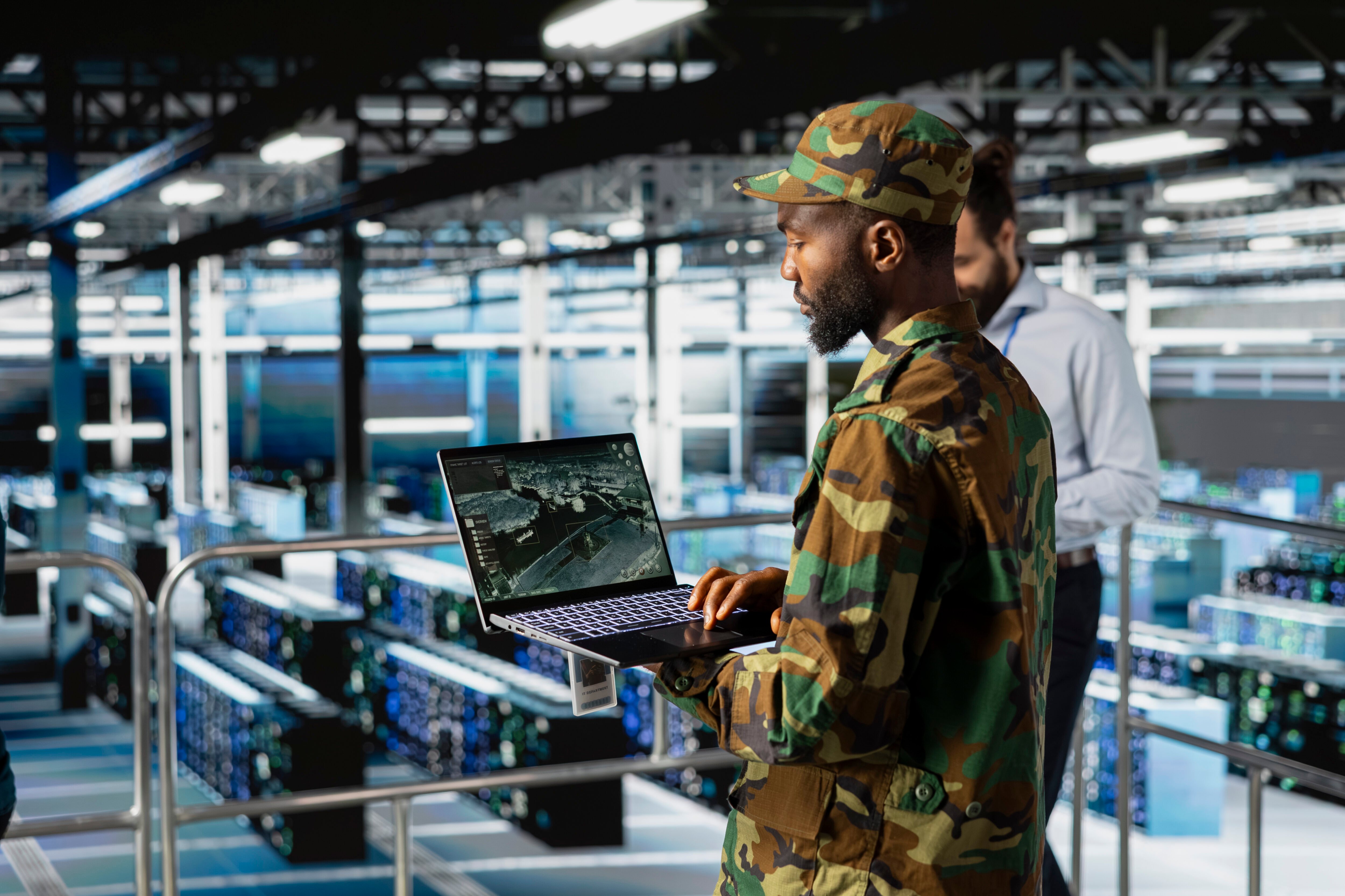 Pessoa em uniforme militar utilizando um laptop em ambiente tecnológico com servidores ao fundo.