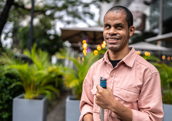 Homem com deficiência visual segurando uma bengala, sorrindo em um ambiente externo com plantas ao fundo.